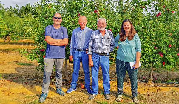 A group of smiling farmers in an apple orchard, holding freshly harvested apples under a bright sky.
