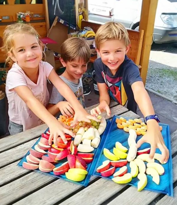 ninos Three happy children reaching for colorful fresh fruit slices, including apples, bananas, and grapes, arranged on trays at an outdoor table.