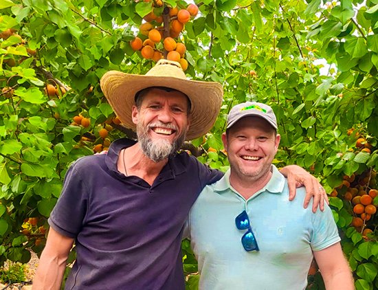 Süße Liebe so groß wie eine Aprikose Two smiling farmers standing in a lush apricot orchard, surrounded by ripe, golden fruit and vibrant green leaves.
