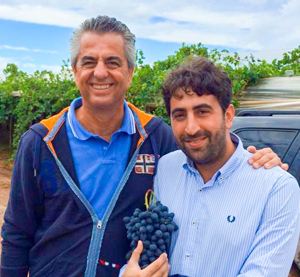 Two smiling men in a vineyard holding fresh black grapes, showcasing premium produce