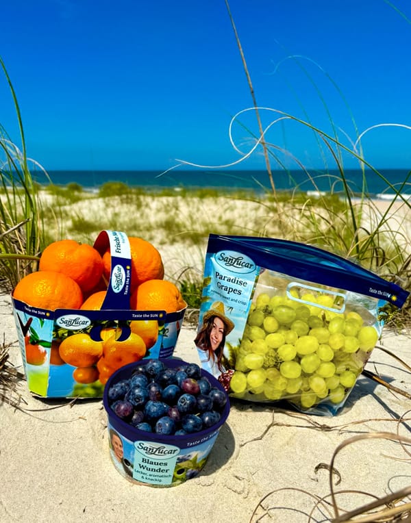 A variety of fresh fruits, including oranges, strawberries, and bananas, displayed on a beach towel with a sunny beach background and clear blue sky.