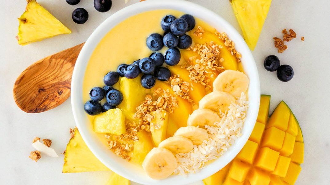 A vibrant smoothie bowl topped with fresh pineapple slices, granola, and seeds, set on a wooden table with a colorful, tropical backdrop.