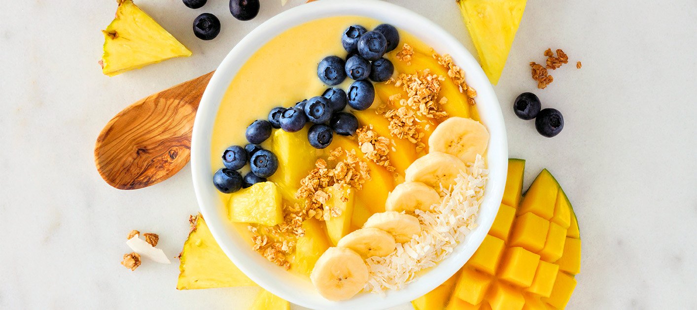 A vibrant smoothie bowl topped with fresh pineapple slices, granola, and seeds, set on a wooden table with a colorful, tropical backdrop.
