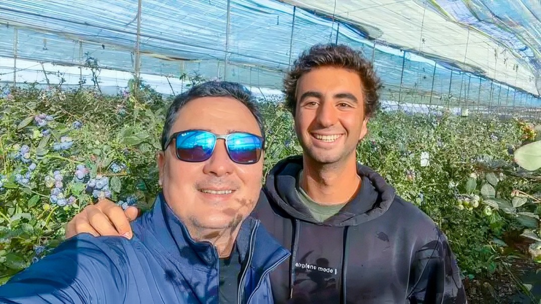 Two smiling men standing together in a blueberry field, surrounded by lush green bushes, with one wearing sunglasses and a jacket, and the other in a hoodie. The blue sky and sunlit background emphasize the fresh, vibrant setting.