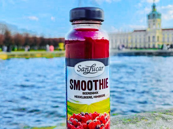 A bottle of SanLucar smoothie, featuring berry flavor with ingredients like strawberries and raspberries, placed on a stone surface near a scenic lake and historical architecture in the background.