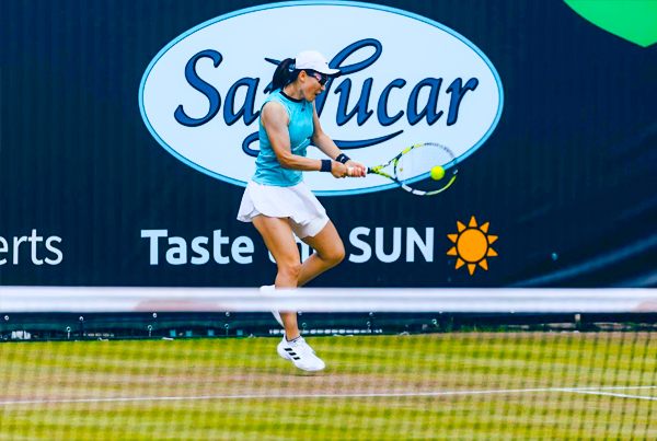 Tenis Female tennis player hitting a backhand shot on a grass court with a large SanLucar "Taste the Sun" banner in the background.