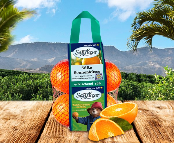 Bag of SanLucar Sonnenküsse oranges with Paddington branding on a rustic table, orchard in background, and sliced oranges.