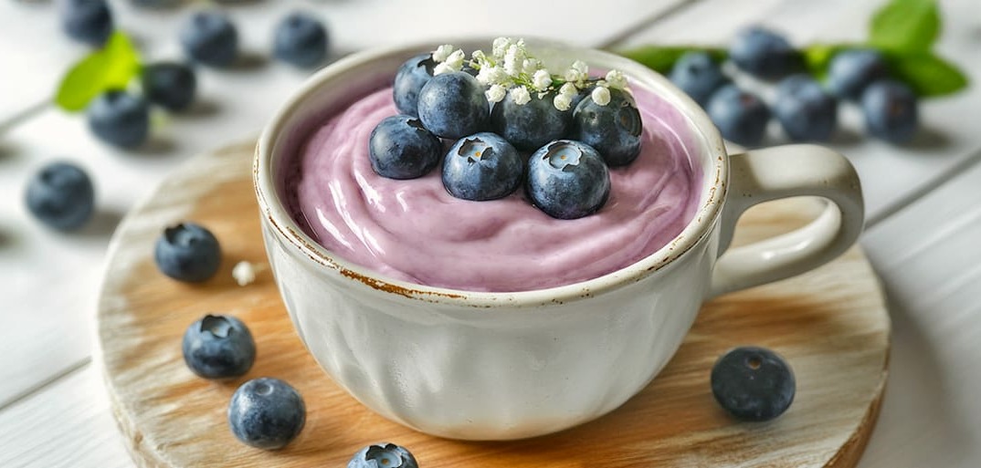 Blueberry Smoothie _Header_2 A cup of creamy blueberry smoothie topped with fresh blueberries and a small flower garnish, placed on a wooden board with scattered blueberries around.