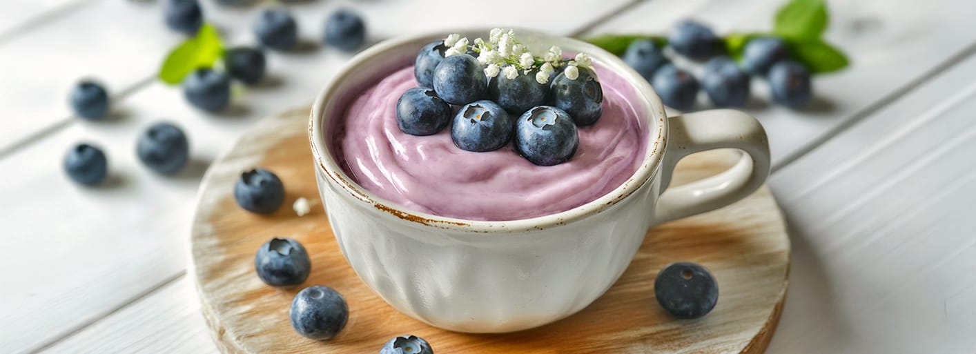 A cup of creamy blueberry smoothie topped with fresh blueberries and a small flower garnish, placed on a wooden board with scattered blueberries around.