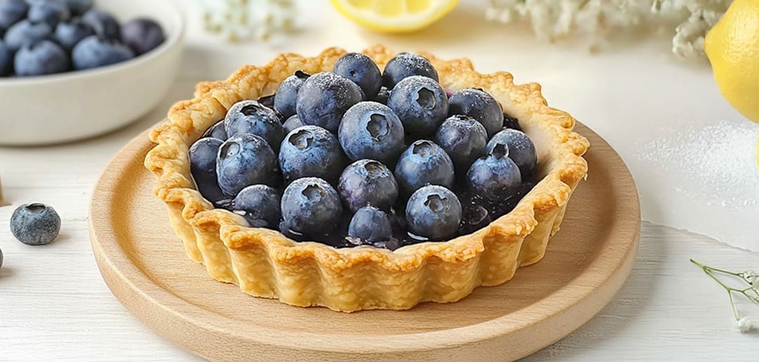 Blueberry tart with a golden crust, topped with fresh, plump blueberries and a hint of lemon, set on a wooden plate.