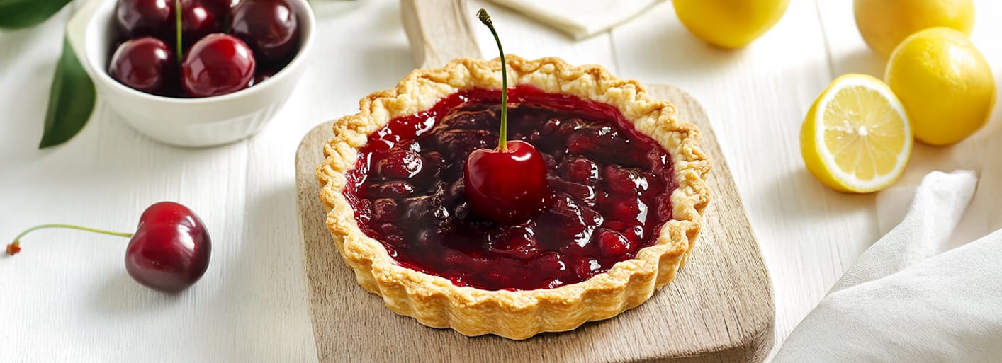 Cherry Tart _Header_3 Cherry tart with lemon and cherries. A full-sized cherry tart topped with a single fresh cherry, with surrounding lemons and a white background.