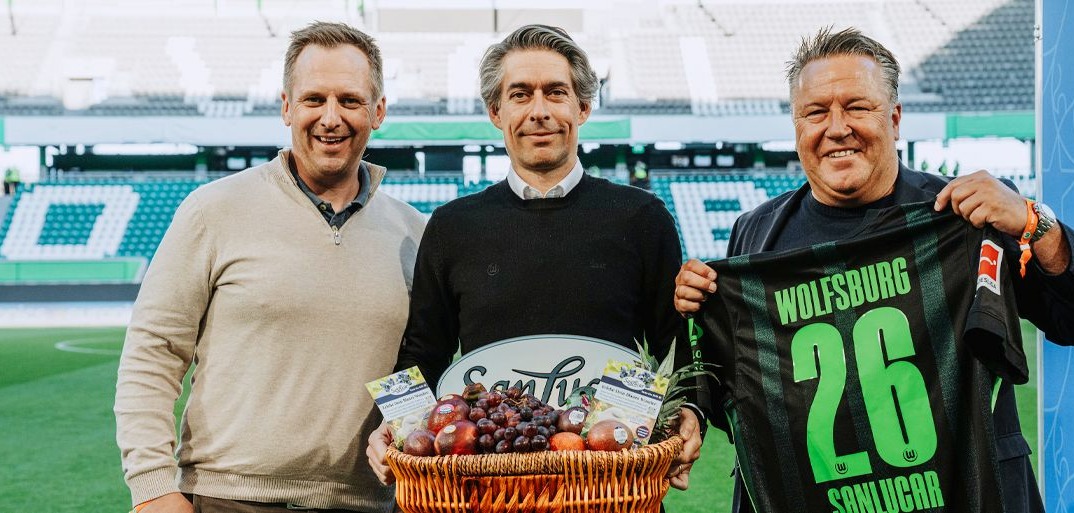 SanLucar partnership event at a football stadium featuring three men, a fruit basket, and a VfL Wolfsburg jersey with number 26.