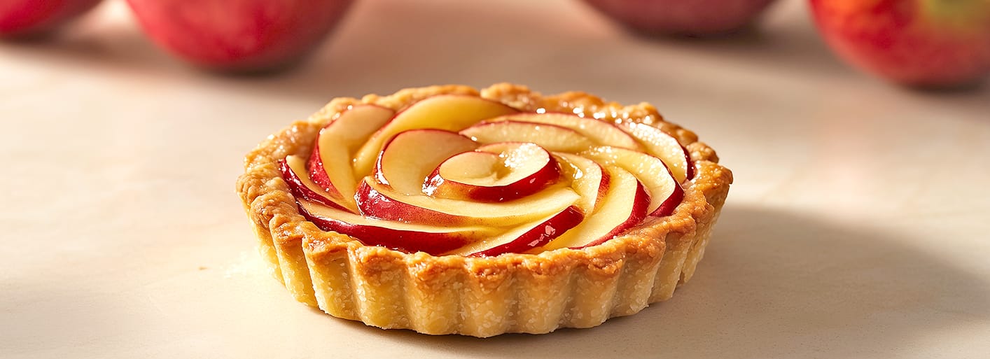 Apple Tart _Header_2 A close-up of a small apple tart with a beautifully arranged spiral of thinly sliced red apples on top. The tart rests on a smooth surface with fresh apples in the background.