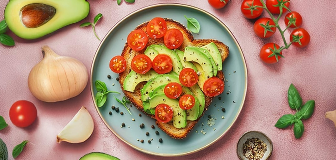 A plate of avocado toast topped with sliced avocado and cherry tomatoes, surrounded by fresh herbs, an avocado, onion, garlic, and seasoning