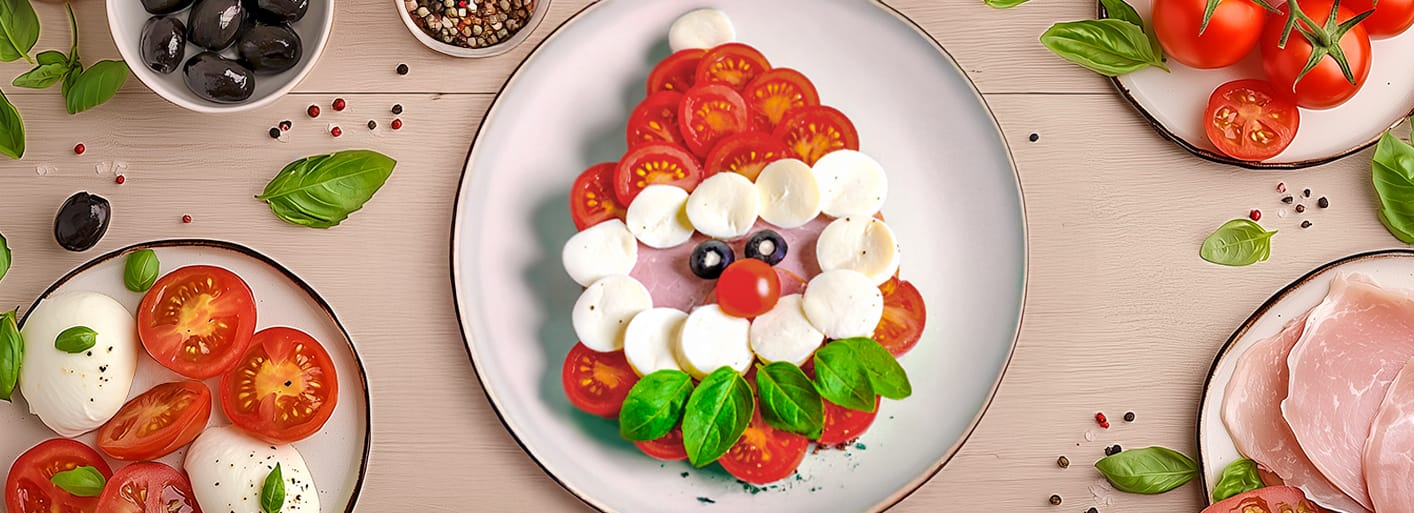 Santalicious Caprese _Header_2 Caprese salad shaped like Santa Claus made with tomatoes, mozzarella, olives, basil, and ham, with a festive setup