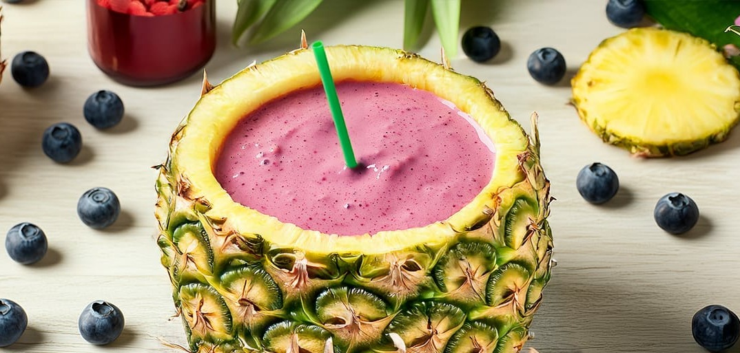 A refreshing summer smoothie served in a hollowed-out pineapple, topped with a colorful blend of berries and a blue straw, surrounded by more fresh fruits and tropical flowers.