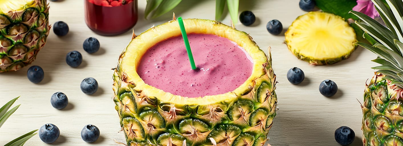 A refreshing summer smoothie served in a hollowed-out pineapple, topped with a colorful blend of berries and a blue straw, surrounded by more fresh fruits and tropical flowers.