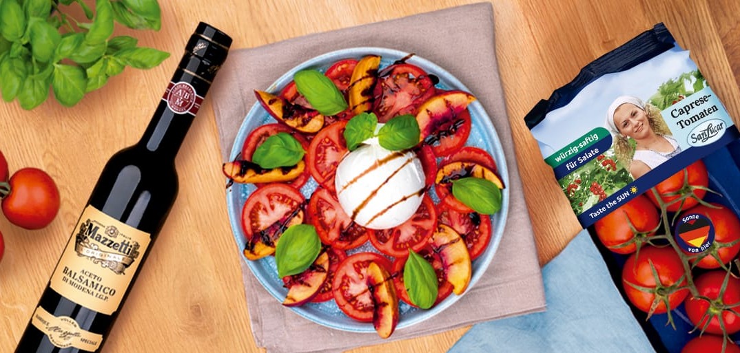 Burrata mit Tomaten und Pfirsichen_Header A colorful salad with fresh tomato and grilled peach slices arranged around a ball of burrata cheese, garnished with fresh basil and drizzled with balsamic vinegar. Next to the plate are vine tomatoes, a bottle of Mazzetti Aceto Balsamico, and a pack of SanLucar cocktail tomatoes.
