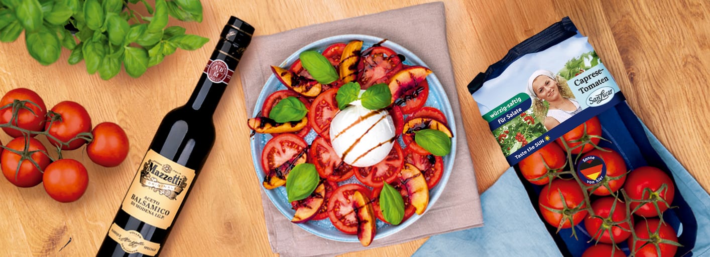 Burrata mit Tomaten und Pfirsichen_Header A colorful salad with fresh tomato and grilled peach slices arranged around a ball of burrata cheese, garnished with fresh basil and drizzled with balsamic vinegar. Next to the plate are vine tomatoes, a bottle of Mazzetti Aceto Balsamico, and a pack of SanLucar cocktail tomatoes.