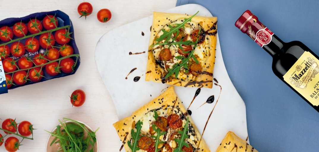 Rectangular puff pastry tarts topped with cherry tomatoes, creamy cheese, fresh arugula, and balsamic glaze on a light serving board. Nearby are a cluster of vine tomatoes, a bottle of Mazzetti balsamic vinegar, and a pack of SanLucar Pralinen-Tomaten tomatoes.