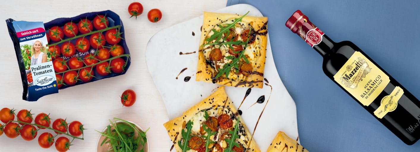 Rectangular puff pastry tarts topped with cherry tomatoes, creamy cheese, fresh arugula, and balsamic glaze on a light serving board. Nearby are a cluster of vine tomatoes, a bottle of Mazzetti balsamic vinegar, and a pack of SanLucar Pralinen-Tomaten tomatoes.