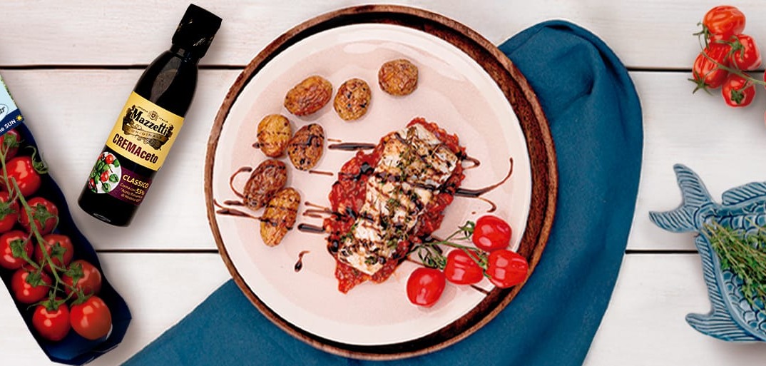 A plate of seared zander fish fillet served over a bed of tomato relish with roasted baby potatoes, drizzled with balsamic glaze. On the side are vine tomatoes and a bottle of Mazzetti CREMAceto balsamic cream along with SanLucar Tomaten-Küsse cherry tomatoes.