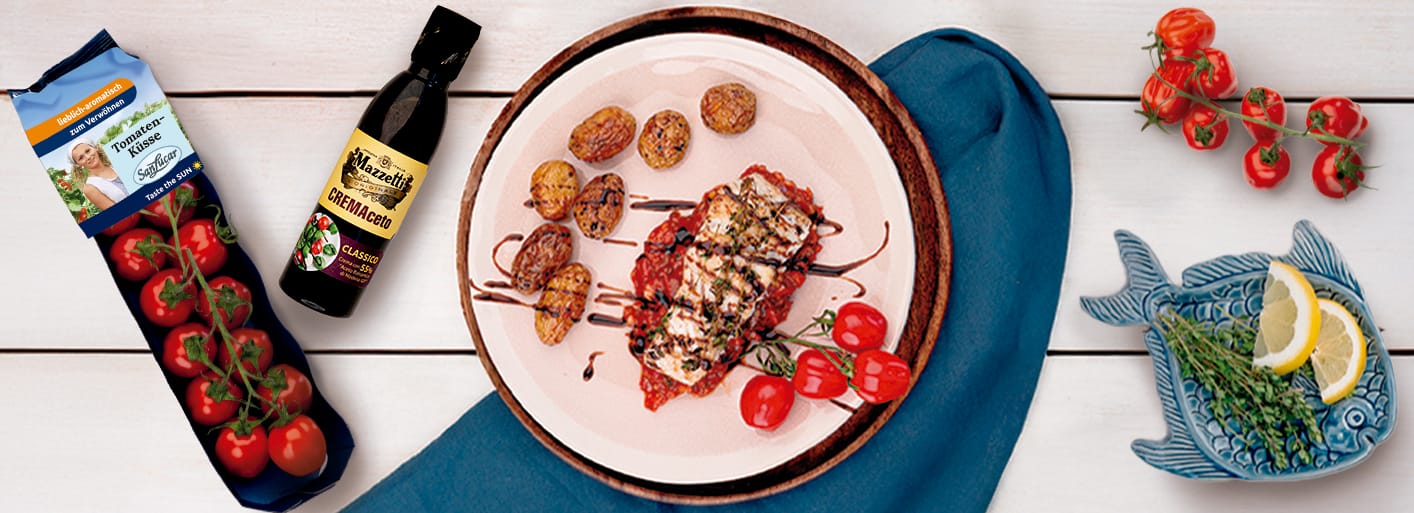 Zander auf Tomaten-Relish mit Balsamico_Header A plate of seared zander fish fillet served over a bed of tomato relish with roasted baby potatoes, drizzled with balsamic glaze. On the side are vine tomatoes and a bottle of Mazzetti CREMAceto balsamic cream along with SanLucar Tomaten-Küsse cherry tomatoes.